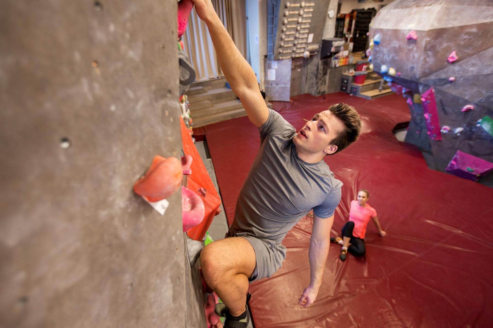 Boulderer zieht sich an einer grauen Wand zum nächsten Griff, unten sitzt eine Frau auf der Matte und schaut zu